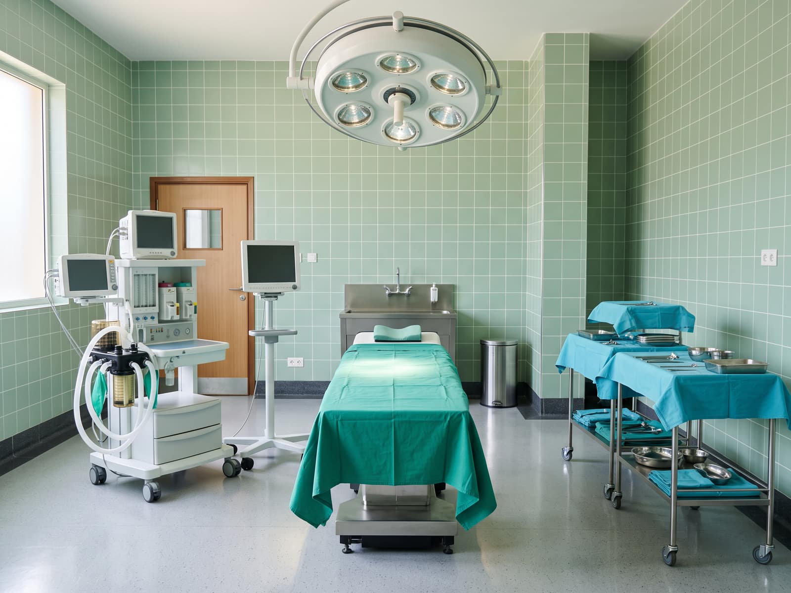 An operating theatre with a table under a round surgical light, an anaesthetic machine and steel trolleys